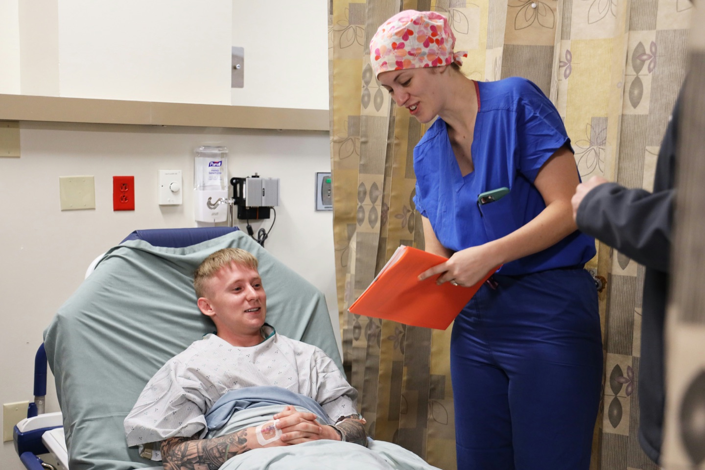 Nurse in blue scrubs speaking with a patient at a hospital bedside